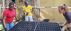Bild zeigt zwei FLINTA, die ein Solarmodul heben, eine erklärende FLINTA steht dabei